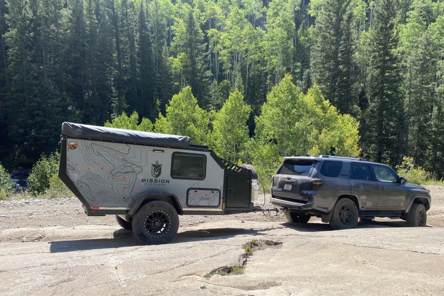This rugged overland RV rental, a trailer towed by an SUV, is set for adventure and off-grid camping amidst a sunny forest.