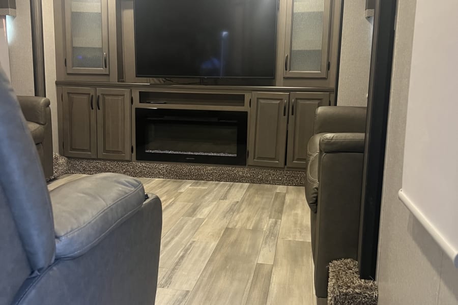 Spacious RV rental living area with large TV, electric fireplace, recliners, and stylish wood-look flooring.