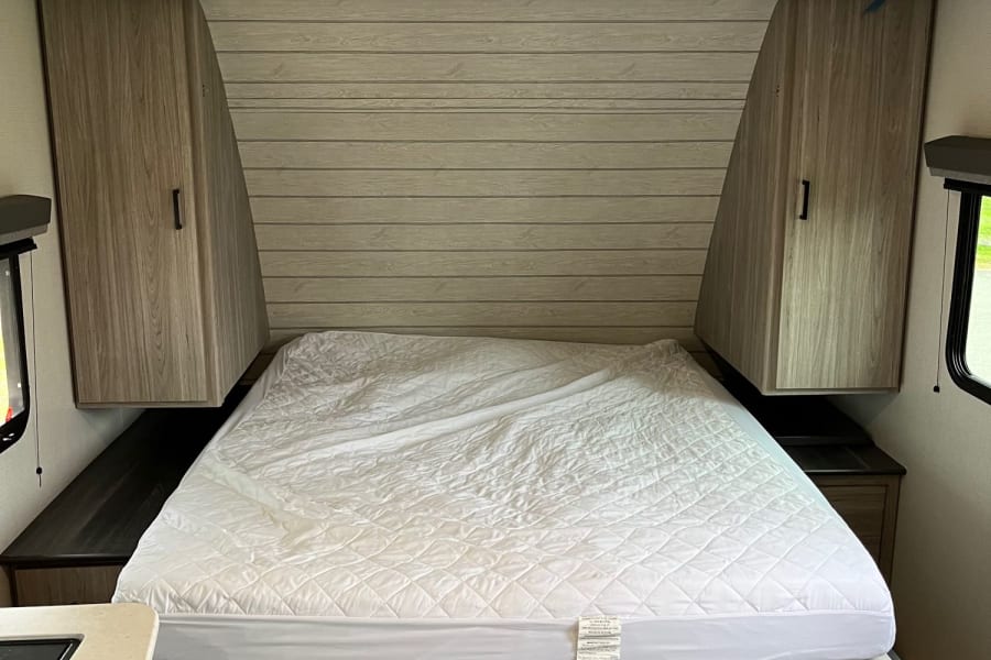 Compact RV rental bedroom with a white quilted bed, angled wood cabinets, and plank-style wall.