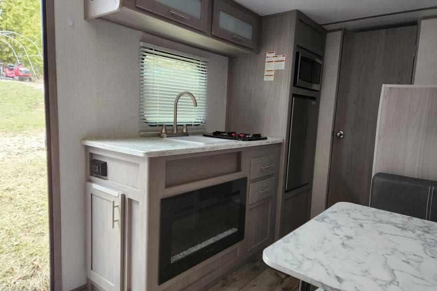 Inside a modern RV rental: a kitchen with sink, stovetop, microwave, fridge, and an electric fireplace for cozy nights.