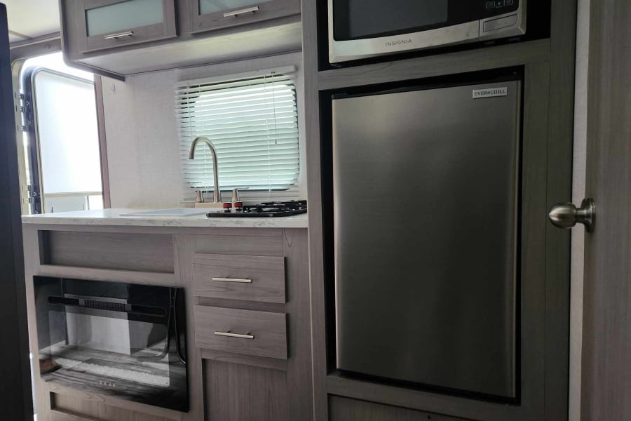 This RV rental's compact kitchen features a sink, stovetop, microwave, refrigerator, and an electric fireplace below.