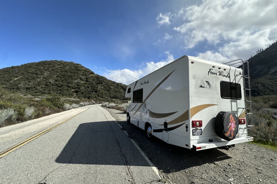 Four Winds RV rental with a Bigfoot spare tire cover, parked on a winding mountain road under a blue sky.