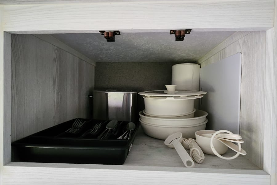 RV rental cabinet stocked with kitchen essentials: toaster, stacked dishes, cutlery tray, and measuring tools.