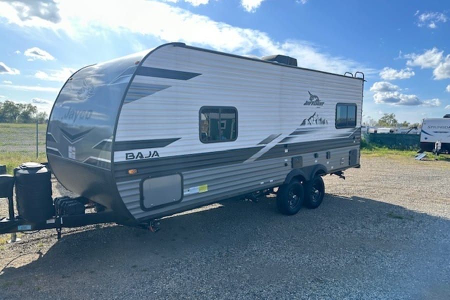 This Jayco Baja travel trailer, a great RV rental option, is parked on gravel under a bright blue sky.
