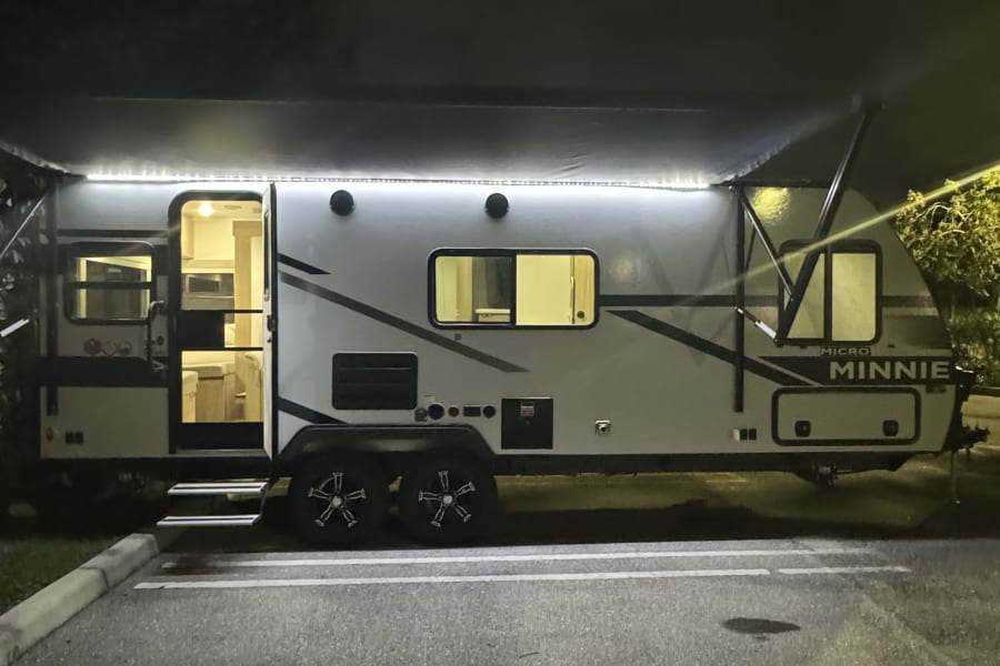 Illuminated Micro Minnie travel trailer RV rental at night, with bright awning lights and an inviting interior visible.