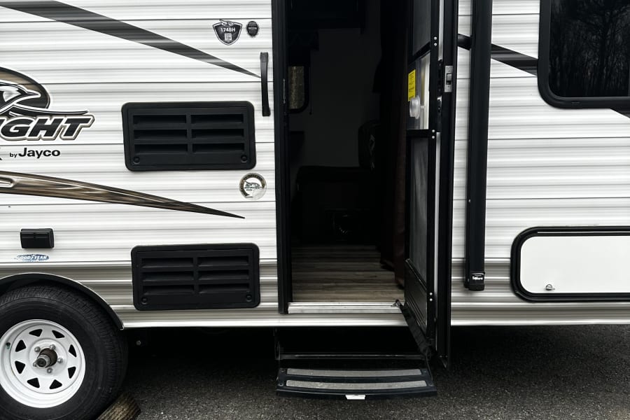 A white Jayco travel trailer, ideal for an RV rental, with its entry door open and steps extended. A tire and wheel chock are visible.