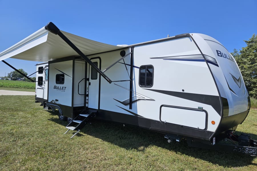 White Bullet Ultra Lite RV rental with extended awning, parked on green grass under a clear blue sky.