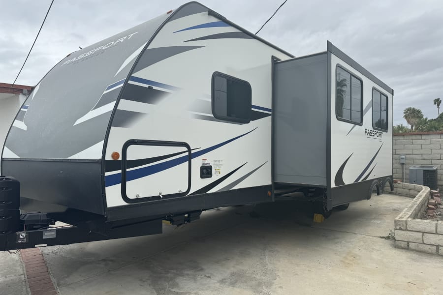 A white, gray, and blue Passport travel trailer **rv rental** with its side slide-out extended, parked on a concrete driveway.