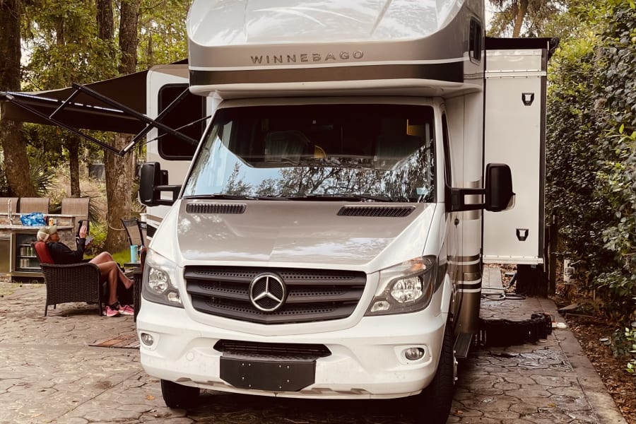 Winnebago RV rental on a paved site. A person relaxes outside the Mercedes-Benz motorhome at this peaceful campsite.