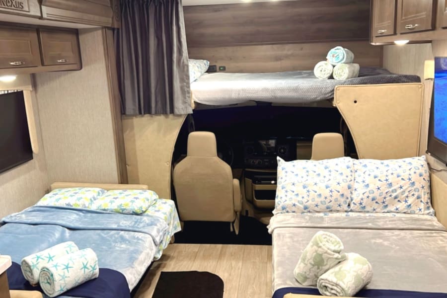 Cozy rv rental interior featuring an overhead bunk bed, two main floor beds with coastal bedding, TV, and driver's cab visible.