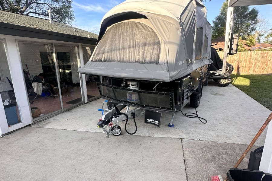 Grey Opus pop-up camper trailer, an RV rental, is fully deployed on a concrete patio beside a house.