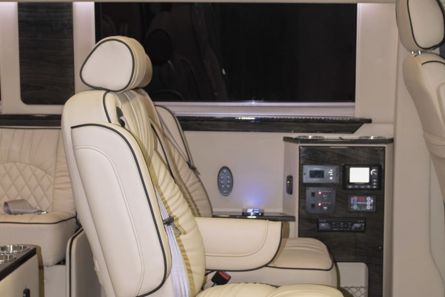 Luxury RV rental interior: Cream leather captain's chairs, dark wood cabinets, TV, and control panel for comfort.