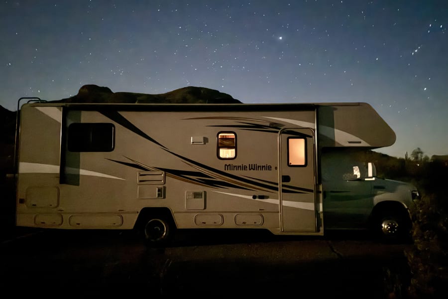 A Winnebago Minnie Winnie RV rental parked under a dazzling star-filled night sky, with mountain silhouettes and glowing interior lights.