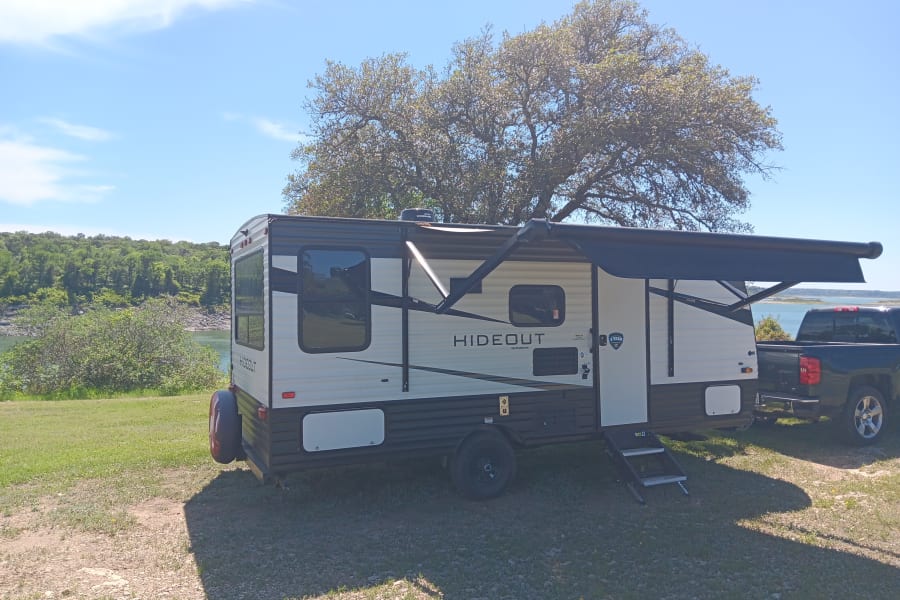 An RV rental (Hideout travel trailer) with an awning extended, parked by a lake, towed by a pickup truck. Perfect for camping!
