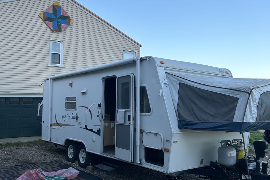 A white Jay Feather hybrid travel trailer, ideal for RV rental, is parked on gravel beside a house with a quilt design.