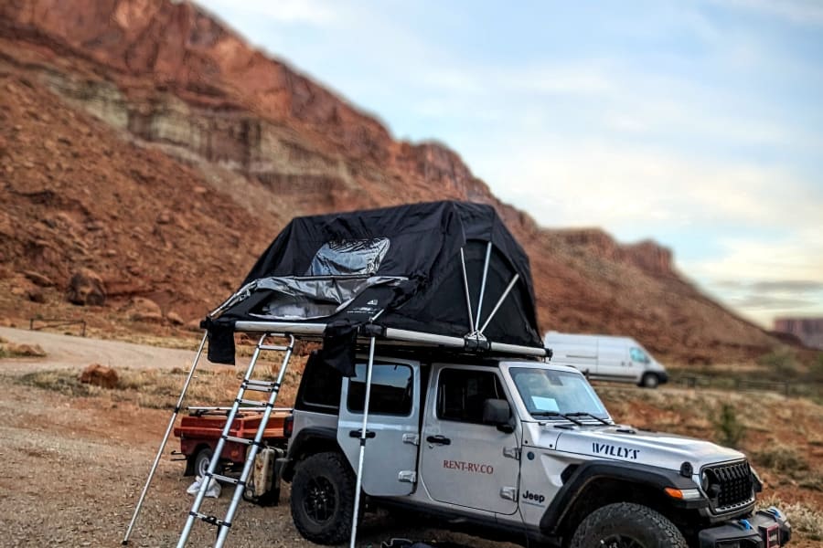 An adventure-ready RV rental Jeep with a rooftop tent set up in a vast desert landscape with red rock formations.