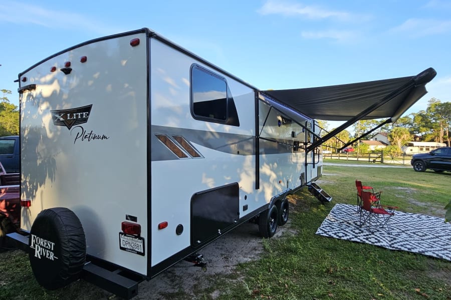 This X Lite Platinum travel trailer rv rental is ready for camping, featuring an extended awning, mat, and red chair.