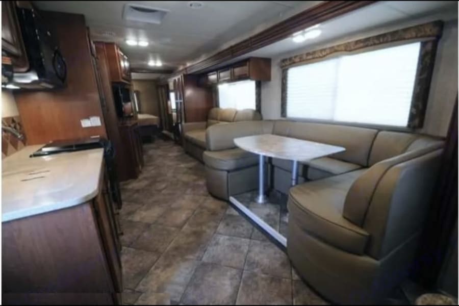 Spacious RV rental interior features a modern kitchen, comfortable dining booth, and sofa. Wood cabinetry & tiled floor.