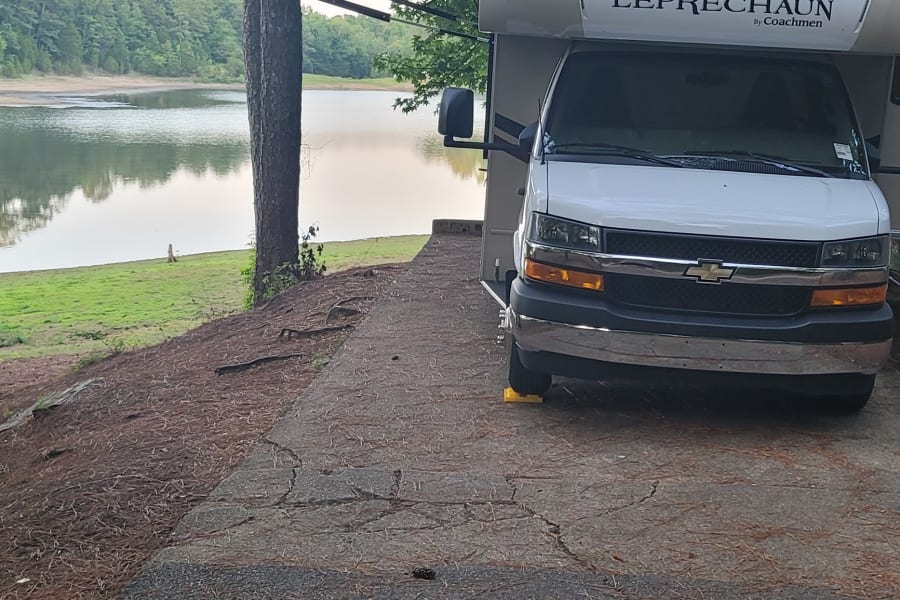 RV rental parked lakeside with awning extended under green trees. Perfect motorhome camping spot.