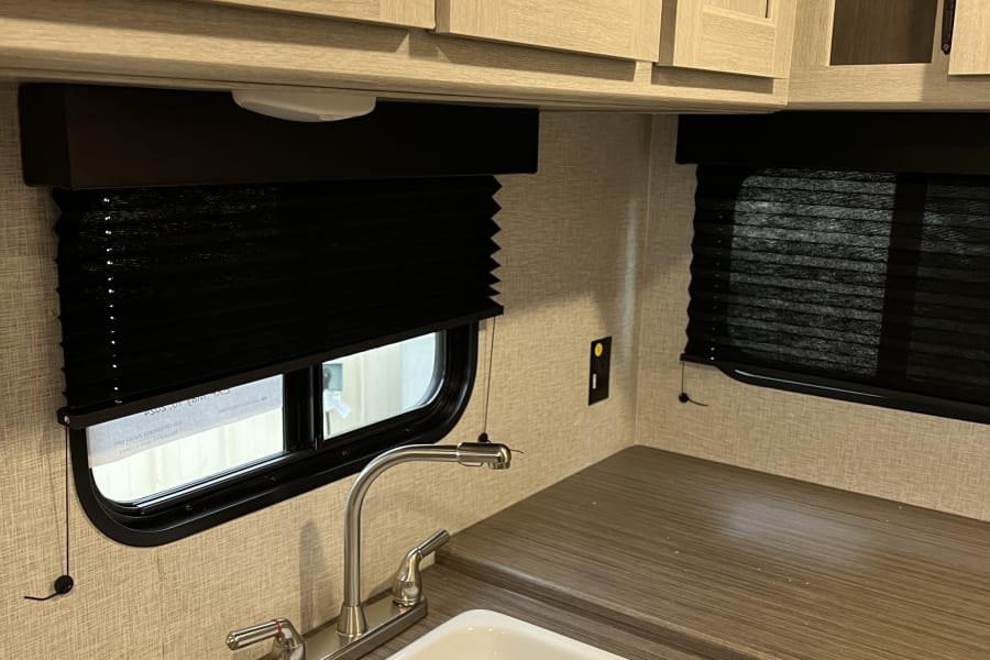RV rental kitchen interior: light wood cabinets, white sink, dark blinds, and counter space.
