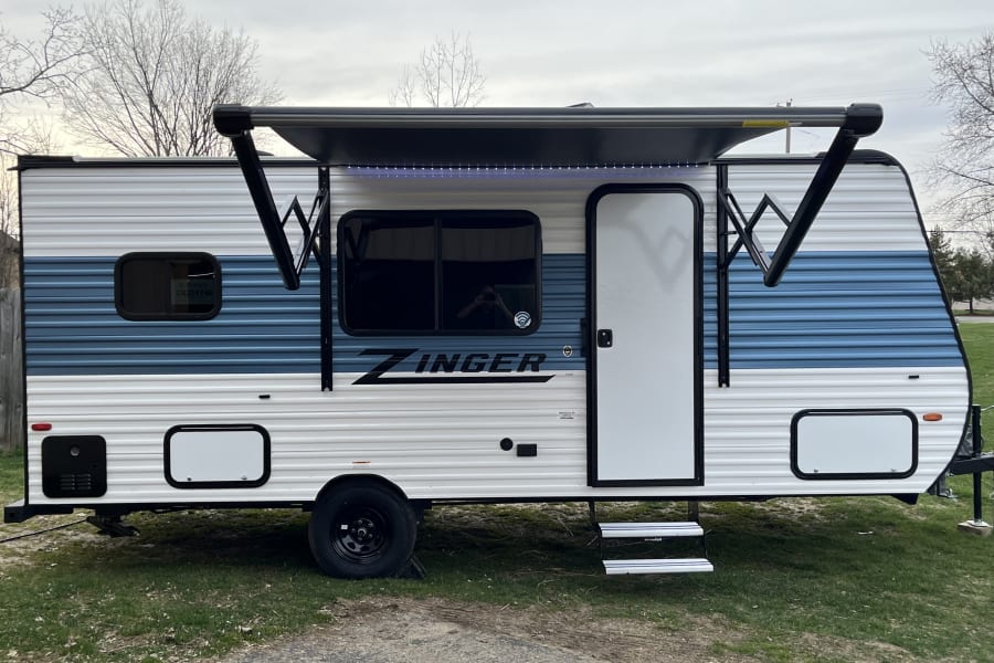 A modern blue and white Zinger travel trailer RV rental, parked with its lighted awning extended.