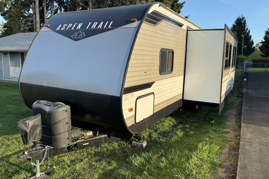 An Aspen Trail travel trailer (RV) with its slide-out extended, parked on sunny green grass. Perfect RV rental.