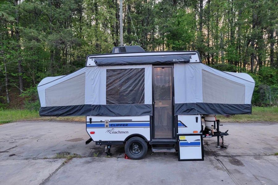 A white and gray Clipper Coachmen pop-up RV rental is fully set up on a paved lot, ready for your camping adventures.