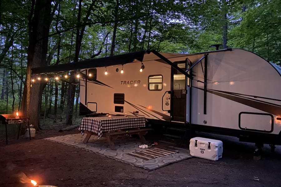 Evening RV rental campsite: cozy travel trailer under string lights, with picnic table, grill, cooler, and campfire.