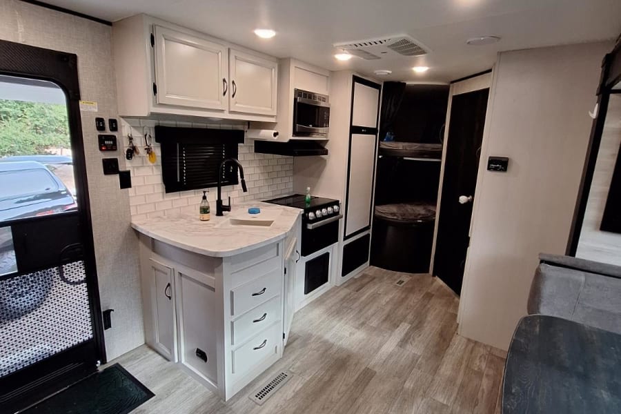 Modern RV rental interior: bright kitchen with white cabinets, light wood-look flooring, and a bunk bed area.