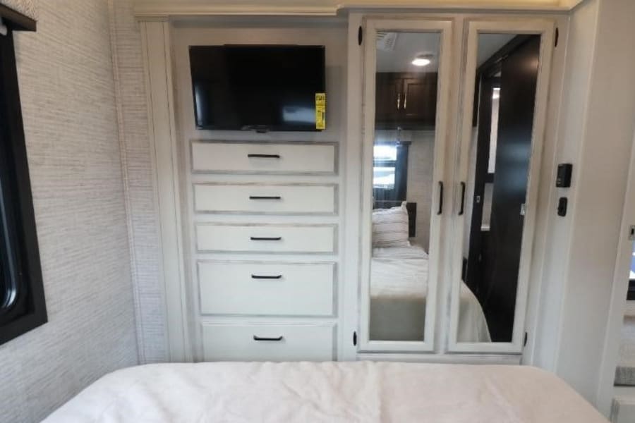 RV rental bedroom: a mounted TV above five white drawers, plus mirrored closet doors reflecting the RV interior. Cozy bed in foreground.