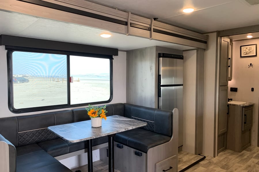 Modern RV rental interior with a cozy dinette overlooking a beach, stainless steel fridge, and storage.