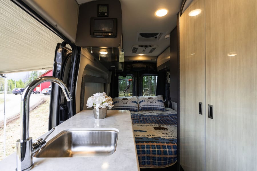 Inside a modern RV rental: compact kitchen with sink, comfy bed, and views of nature through the open door.