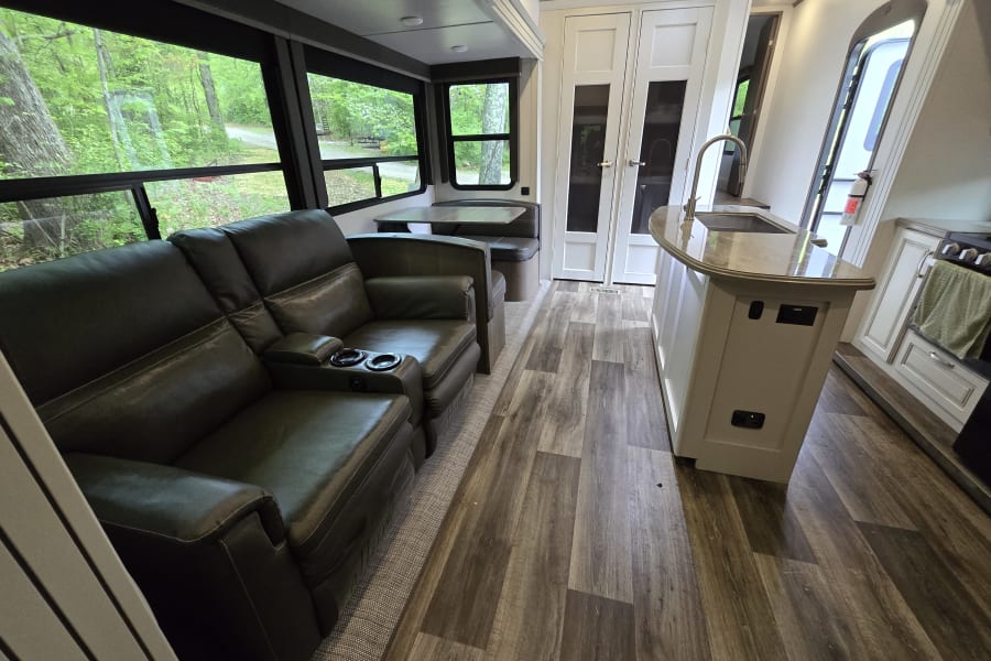 Spacious RV rental interior with comfy recliners, a dinette, and kitchen. Large windows offer serene views of nature outside.