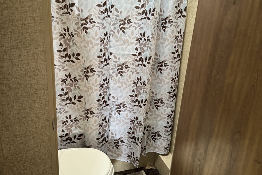 RV rental bathroom interior features a white toilet, leaf-pattern shower curtain, tile floor, rug, and waste bin.