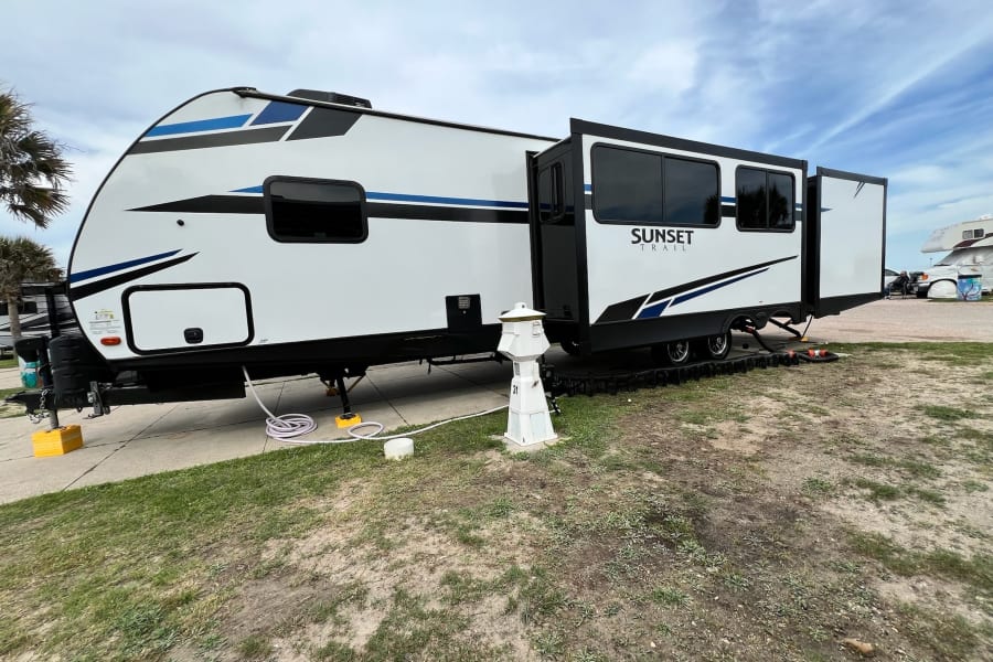 A modern white and blue Sunset Trail RV rental is fully set up at a spacious campsite, its slide-out extended.