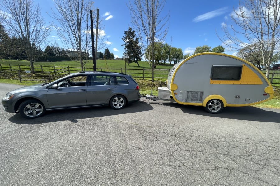 Grey VW station wagon towing a yellow/grey teardrop camper on an asphalt road. Perfect RV rental for your next adventure.