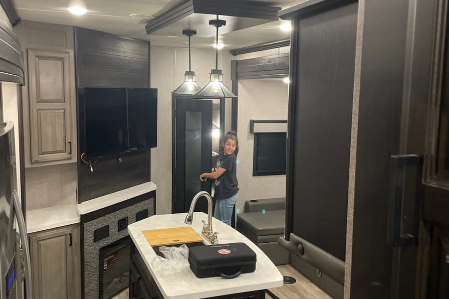 Inside an RV rental: a modern interior with kitchen island, living space, and a girl by the sink. Ideal for travel.