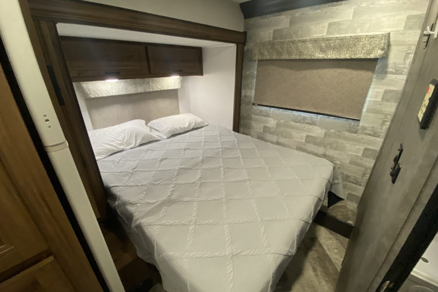 Compact RV rental bedroom showing a bed with white linens, overhead cabinets, and a window with a pulled shade.