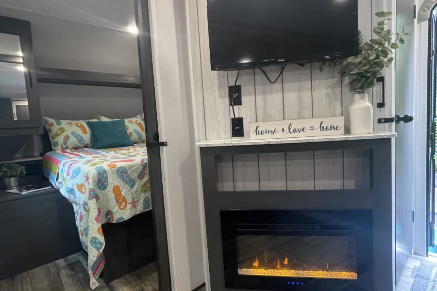 Modern RV rental interior: bedroom with colorful bedding, living area with TV & electric fireplace, wood-look floor.
