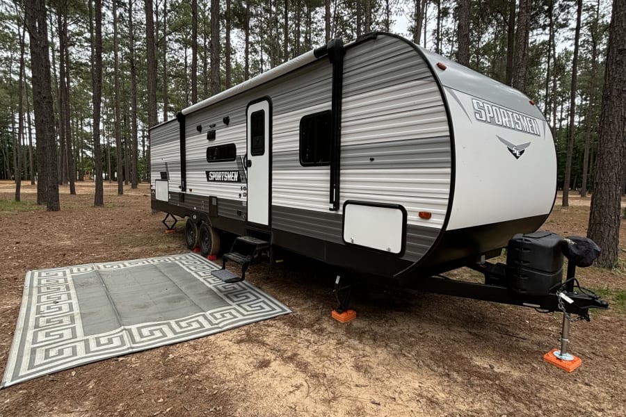 An RV rental is parked with an outdoor rug among tall pine trees at a tranquil campsite, ready for your adventure.