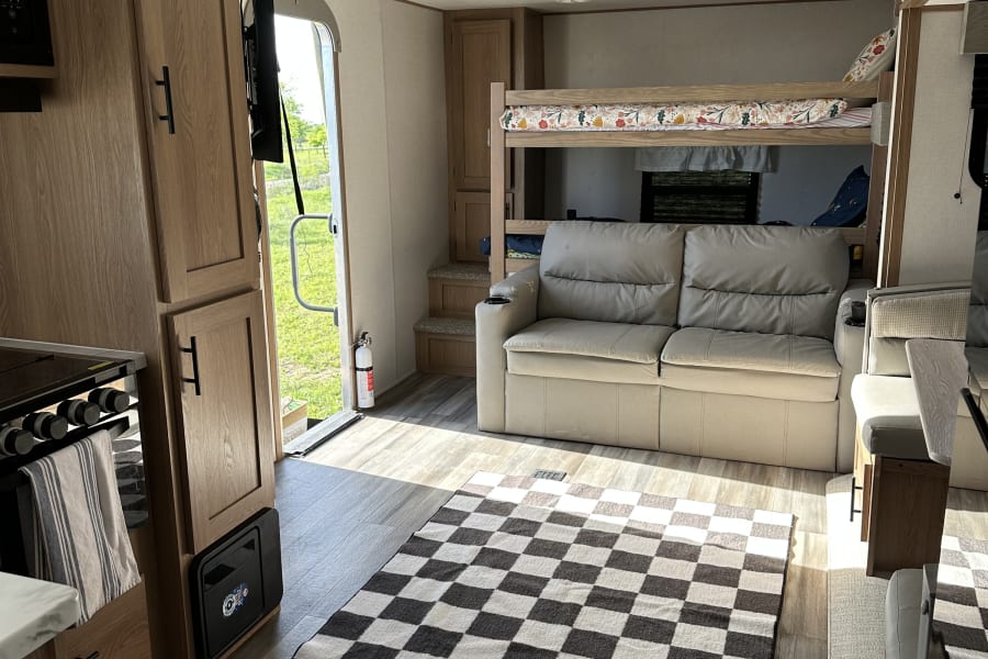 Bright RV rental interior with comfy sofa, kitchen, bunk beds, checkered rug, and a peek outside.
