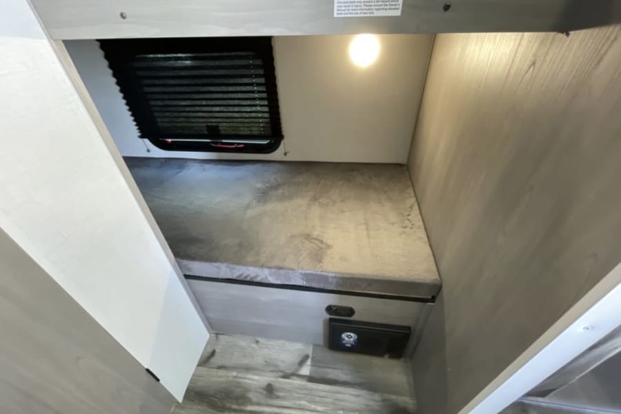 Compact RV rental lower bunk features a cozy gray mattress, window with blinds, overhead light, and unit controls below.