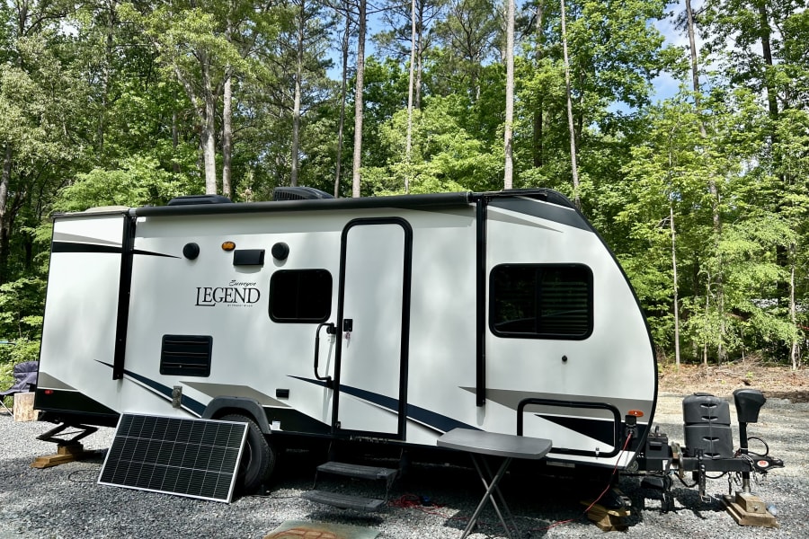 A white 'Legend' RV rental is parked on a gravel site amidst tall trees, with a solar panel and small table setup.