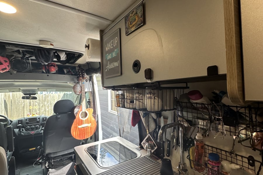 Inside an RV rental, showing the cozy kitchen with sink, stovetop, and clever storage solutions. Guitar hangs near the cab.