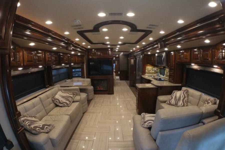 This luxury RV rental showcases a spacious interior with light sofas, dining, fireplace, and a full kitchen.