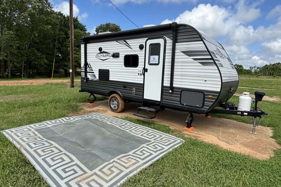 An Olympia Sport RV rental is set up at a rural campsite, with an ornate outdoor rug on grass.