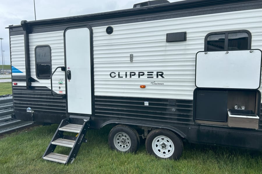 This Coachmen Clipper travel trailer, an ideal RV rental, features a white & black exterior, outdoor kitchen with fridge/sink, and accessible steps.