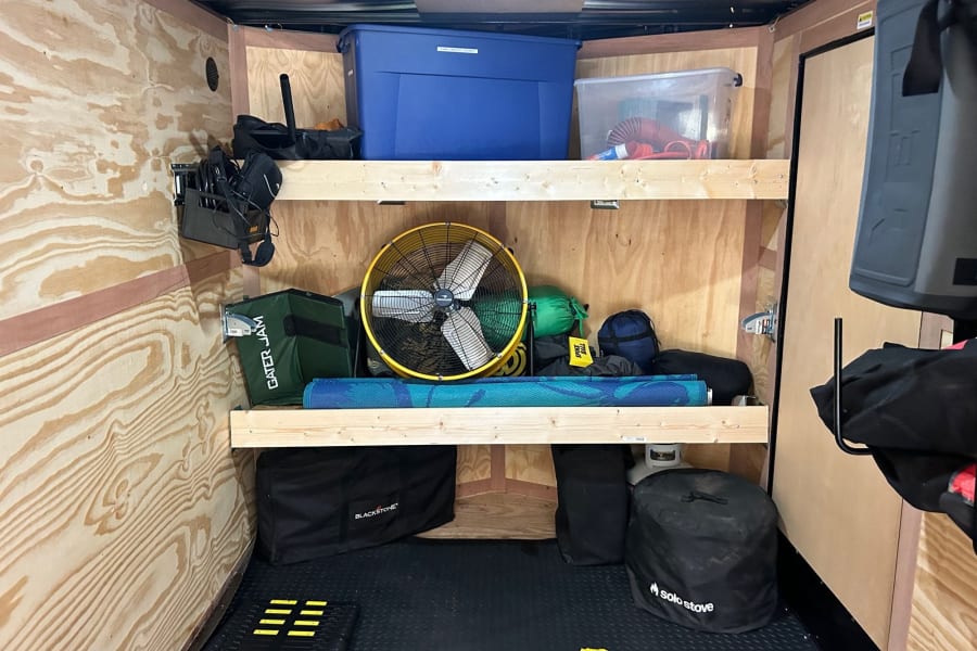 Inside an **rv rental**: organized storage area with wooden shelves, bins, and outdoor gear like a fan, mat, and stove.