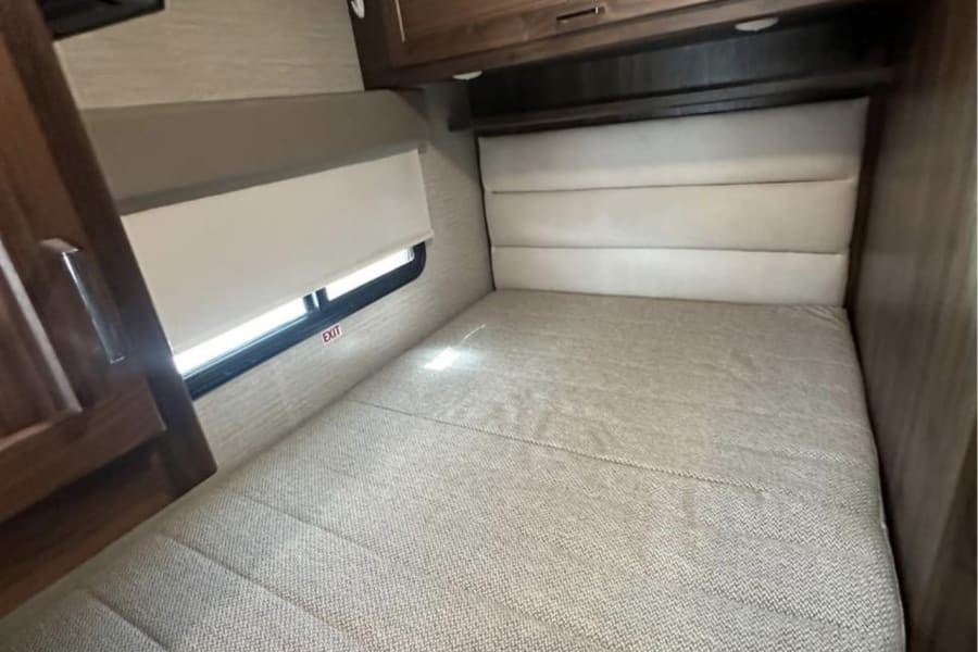 RV rental bedroom featuring a spacious bed with padded headboard, window with blind, and overhead cabinets for storage.
