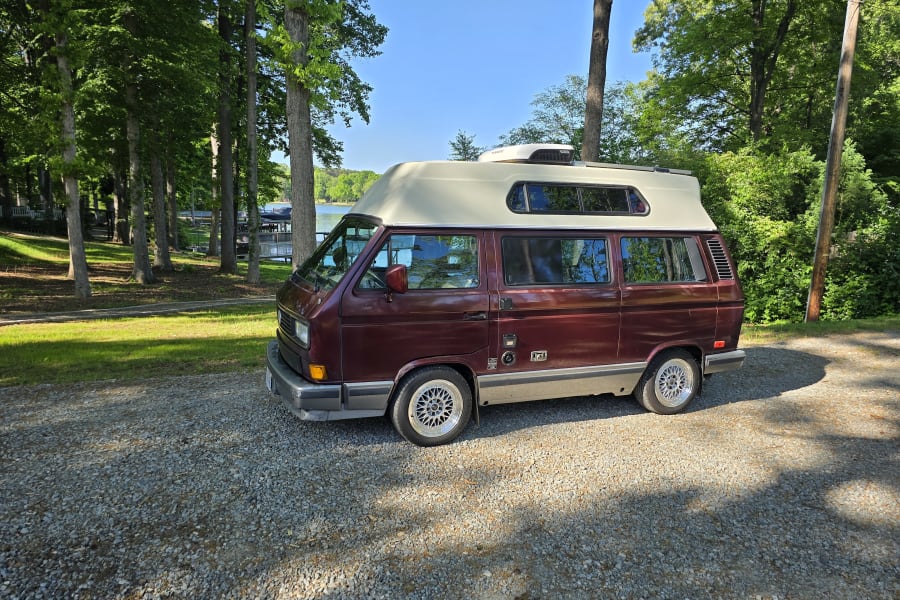 Maroon VW Vanagon camper van on gravel by a lake and trees, perfect for your next RV rental adventure.